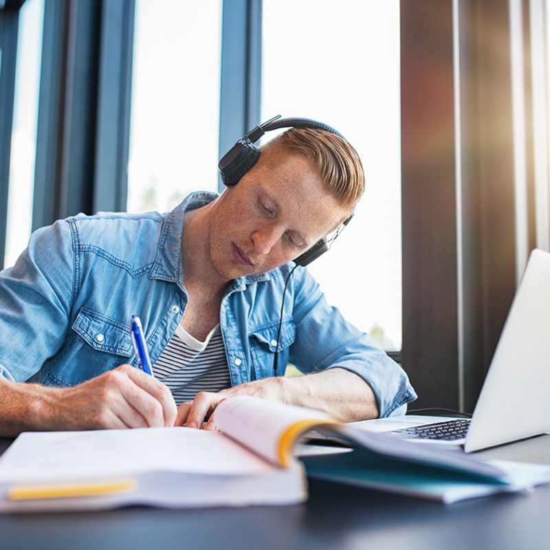 Shot of young man studying in college library sitting at table with laptop and books. Male university student preparing notes for the exam at library.